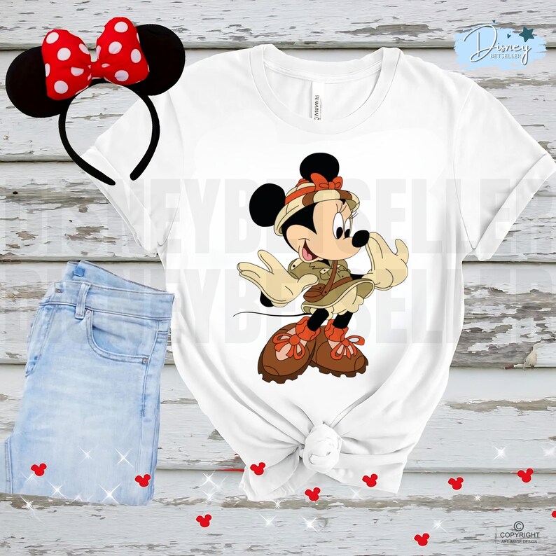 May include: A white t-shirt featuring a graphic of Minnie Mouse dressed in a tan safari outfit with a matching hat and brown boots. Beside the shirt is a black headband with a red and white polka dot bow and black mouse ears.