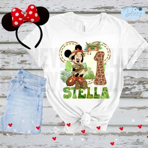May include: White t-shirt with a Minnie Mouse design, the name "STELLA", and the number "1". The design has a safari theme with a giraffe print and tropical leaves. A red and white polka dot bow headband and blue jeans are also in the image.