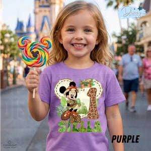 May include: A girl smiles, holding a Mickey Mouse-shaped lollipop. She wears a purple t-shirt with a Minnie Mouse safari design, the number 1, and the name "Stella". The word "PURPLE" is printed at the bottom.