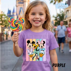 May include: A purple t-shirt featuring a colorful graphic of Disney characters, including Mickey Mouse, Minnie Mouse, Donald Duck, and Goofy. The shirt has vertical pastel stripes and the word "PURPLE" in the bottom right corner. A child is holding a colorful Mickey Mouse lollipop.
