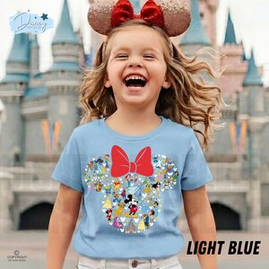 May include: Light blue t-shirt featuring a Minnie Mouse design with a red bow and various Disney characters. The shirt has the text "LIGHT BLUE" at the bottom. The image shows a young girl wearing the shirt and a Minnie Mouse ear headband.