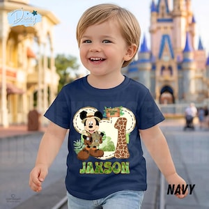 May include: Navy blue toddler t-shirt with a Mickey Mouse safari design, the number 1, and the name "Jaxson". The design incorporates a giraffe print and tropical leaves. The shirt is labelled "Navy".