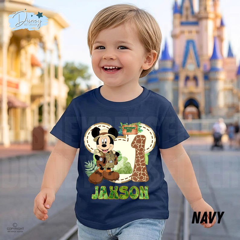 May include: Navy blue toddler t-shirt with a Mickey Mouse safari design, the number 1, and the name "Jaxson". The design incorporates a giraffe print and tropical leaves. The shirt is labelled "Navy".