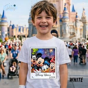 Puede incluir: Una camiseta blanca con un dise&ntilde;o tipo polaroid de los personajes de Disney, Mickey y Minnie Mouse, Donald Duck y Goofy, con el texto "Disney World 2026". La camiseta la lleva un ni&ntilde;o sonriente, con un parque tem&aacute;tico de fondo.