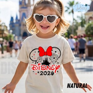 May include: A light beige t-shirt featuring a Disney design with a red bow, fireworks, and a castle silhouette. The word "Disney" is in red, with "2026" below. The shirt is labeled "NATURAL" in black text.