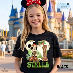May include: Black t-shirt with a Minnie Mouse design, the text "Birthday Girl", and the name "Stella". The design includes a giraffe print number 1 and tropical foliage. The girl is wearing a Minnie Mouse ear headband.