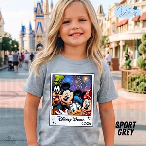 Puede incluir: Una camiseta gris claro con un gr&aacute;fico de Mickey Mouse, Minnie Mouse, Donald Duck y Goofy con fuegos artificiales y el texto "Disney World 2026". La camiseta la lleva una ni&ntilde;a peque&ntilde;a con el pelo rubio.