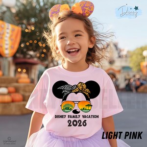 May include: A light pink t-shirt featuring a Minnie Mouse design with sunglasses and the text "Disney Family Vacation 2026". The shirt is worn by a child with a matching headband.