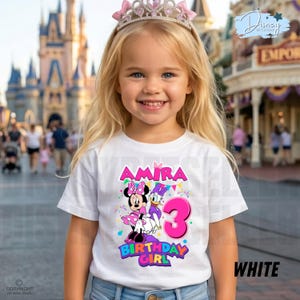 May include: A white short-sleeved t-shirt featuring a colorful graphic of Minnie Mouse and Daisy Duck, the name Amira, and a large pink number 3. The design includes festive confetti and the text Birthday Girl in bright, bold, multi-colored lettering.