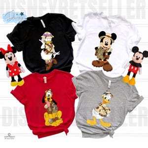 May include: Four short-sleeve t-shirts in black, white, red, and gray. The black shirt features Daisy Duck in safari gear, the white shirt shows Mickey Mouse in safari attire, the red shirt displays Pluto, and the gray shirt shows Donald Duck.