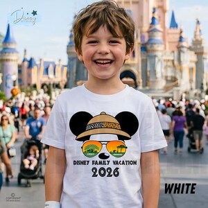 May include: A white t-shirt featuring a Mickey Mouse design wearing a safari hat and sunglasses with the text "Disney Family Vacation 2026". The shirt is worn by a smiling child in front of a theme park.