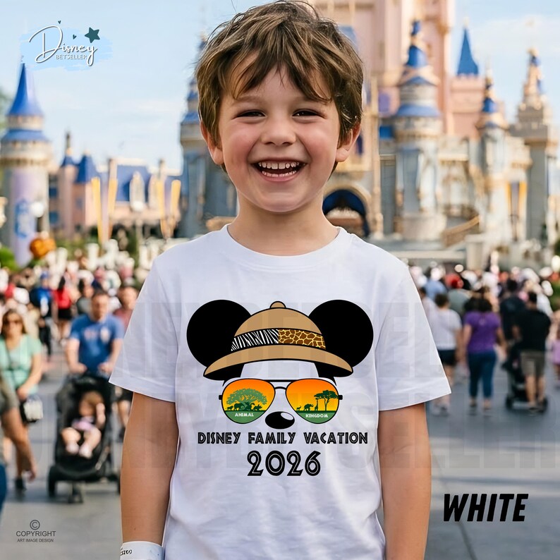May include: A white t-shirt featuring a Mickey Mouse design wearing a safari hat and sunglasses with the text "Disney Family Vacation 2026". The shirt is worn by a smiling child in front of a theme park.