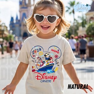 May include: A cream-coloured t-shirt featuring a graphic of Mickey Mouse, Minnie Mouse, Donald Duck, and Goofy on a cruise ship inside a Mickey Mouse ear silhouette. The design includes the text 2026, NAME, and Disney Cruise in red and blue.