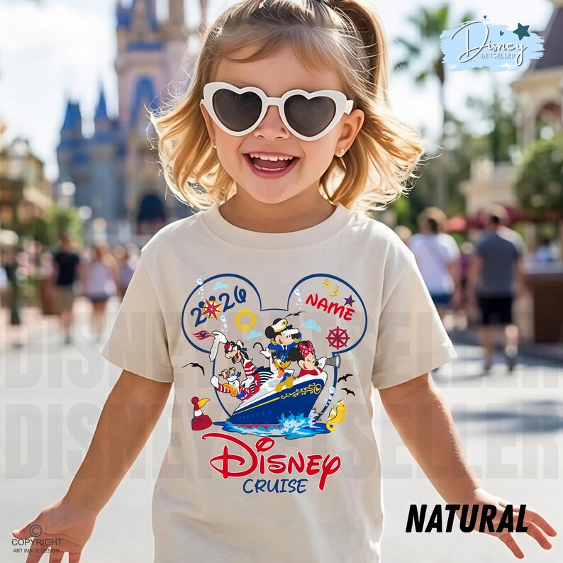 May include: A cream-coloured t-shirt featuring a graphic of Mickey Mouse, Minnie Mouse, Donald Duck, and Goofy on a cruise ship inside a Mickey Mouse ear silhouette. The design includes the text 2026, NAME, and Disney Cruise in red and blue.