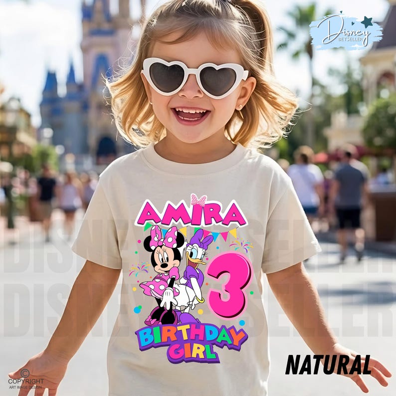 May include: A beige short-sleeved t-shirt featuring a graphic of Minnie Mouse and Daisy Duck, the name AMIRA in pink, the number 3 in large pink font, and the text BIRTHDAY GIRL in colorful letters, with festive bunting and confetti accents.
