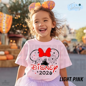 May include: A light pink t-shirt with a Disney graphic featuring Minnie Mouse ears, a red bow, fireworks, and a castle. The text "Disney 2026" is printed below the graphic. The girl is wearing a purple tutu and a headband with Mickey Mouse ears.