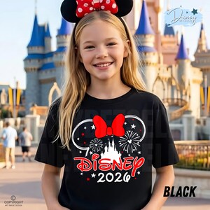May include: A black t-shirt featuring a Minnie Mouse design with a red bow, fireworks, and the word "Disney" in red script. The year "2026" is printed below. The shirt is worn by a person wearing a Minnie Mouse ear headband.