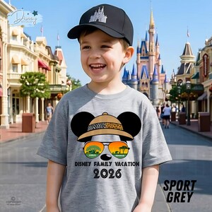 May include: A gray t-shirt with a Mickey Mouse design wearing a safari hat and sunglasses, with the text "Disney Family Vacation 2026." The shirt is paired with a black baseball cap featuring a castle graphic. The words "Sport Grey" are printed on the shirt.
