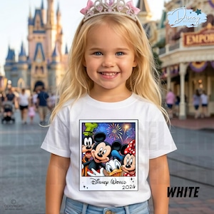 Puede incluir: Una ni&ntilde;a peque&ntilde;a con una camiseta blanca con un gr&aacute;fico colorido de personajes de Disney, incluyendo Mickey Mouse, Minnie Mouse, Donald Duck y Goofy, con fuegos artificiales y el texto "Disney World 2026". Tambi&eacute;n lleva una tiara rosa.