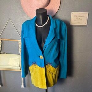 Op de afbeelding: Een turquoise blazer met een gele suède onderpaneel en geborduurde details. De blazer heeft een enkele gouden knoop en wordt gepresenteerd op een zwarte mannequin. Een roze zonnehoed is op de achtergrond.