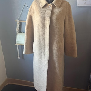 May include: A long, beige, textured coat with a classic collar, long sleeves, and a single visible button closure at the neck. The coat features a straight silhouette, side pockets, and a soft, fuzzy exterior finish, displayed on a mannequin.