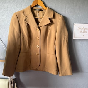 Op de afbeelding: Een beige blazer met een reverskraag, drie knopen en twee voorzakken. De blazer hangt aan een houten kleerhanger. De merknaam "Marks & Spencer" is zichtbaar op het label.