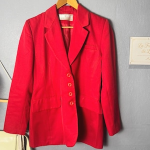 Peut inclure: Un blazer rouge vif doté d'un revers cranté, de manches longues et d'une fermeture frontale à quatre boutons. La veste comprend deux poches à rabat sur le devant et une poche poitrine, avec une étiquette de marque blanche au col sur un cintre.