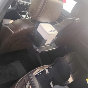 May include: A car interior featuring brown leather seats and a black headrest-mounted organiser. The organiser holds a white container with a lid and a small tray. A child car seat is visible in the back seat.