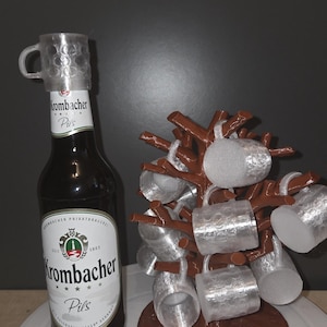 May include: A brown glass Krombacher Pils beer bottle with a clear plastic cap topper, positioned next to a brown tree-shaped stand holding several small, clear, textured plastic beer mugs hanging from its branches.