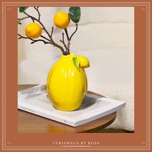 Peut inclure: Un vase en céramique jaune brillant en forme de citron, contenant une branche avec trois citrons artificiels et des feuilles vertes. Le vase est posé sur un livre blanc avec le texte "Southern France" sur une table en bois. Un élément de décoration intérieure.