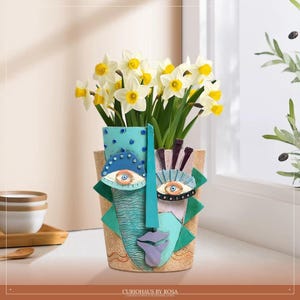 May include: A decorative face-shaped vase with a teal nose and lips, and two eye designs. The vase is filled with yellow and white daffodils. The vase has a textured, tan background with green and blue accents. The text "CURIOHAUS BY ROSA" is at the bottom.