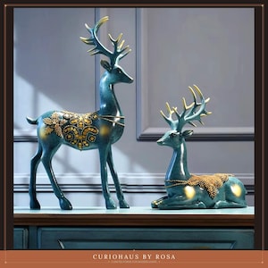 May include: Two teal deer figurines with gold-coloured antlers and intricate gold-coloured decorative patterns on their backs. One deer is standing upright while the other is in a reclining position. The text CURIOHAUS BY ROSA is at the bottom.