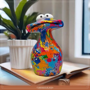 Peut inclure: Un vase en céramique coloré et fantaisiste avec un design unique en forme de canard. Le vase est recouvert d'éclaboussures de peinture vibrantes bleues, oranges, jaunes et roses. Il a de grands yeux blancs et une large bouche ouverte. La base est arrondie.