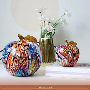 May include: Two apple-shaped sculptures featuring a vibrant, multi-colored graffiti pattern with bold orange, blue, pink, and black lines. Each apple is topped with a metallic gold stem and leaf, creating a striking contrast against the colorful surface.