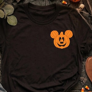 May include: A black short-sleeve t-shirt featuring a small orange graphic on the left chest area. The graphic depicts a pumpkin shaped like the silhouette of a mouse head with a smiling face. The shirt is displayed alongside blue denim jeans.