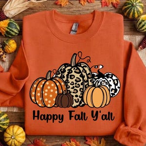 May include: An orange crewneck sweatshirt featuring a graphic of four pumpkins with varying patterns, including polka dots and leopard print, alongside the black text "Happy Fall Y'all" printed across the center of the garment.