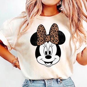 Leopard print Minnie Mouse head with sparkling galaxy-style black ears and a cheetah print bow, Disney-inspired graphic design on a transparent background. Cute Disney vacation shirt design for women, girls, and matching family trip tees