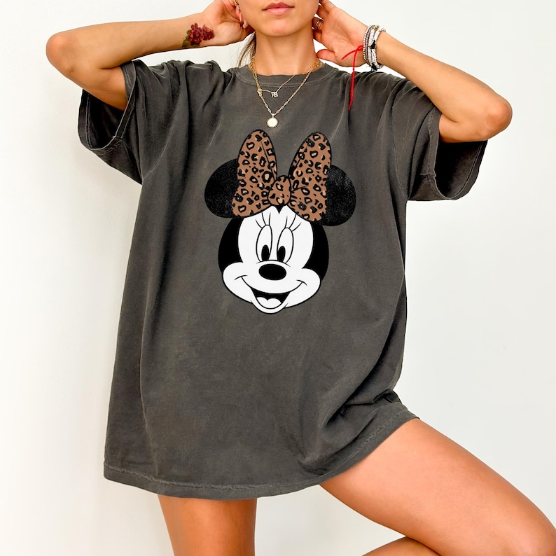 Leopard print Minnie Mouse head with sparkling galaxy-style black ears and a cheetah print bow, Disney-inspired graphic design on a transparent background. Cute Disney vacation shirt design for women, girls, and matching family trip tees