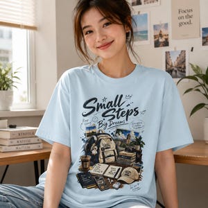 Könnte beinhalten: Ein hellblaues T-Shirt mit einem Grafikdruck und dem Text "Small Steps Big Dreams". Das Design zeigt Illustrationen eines Rucksacks, Büchern, einer Kamera, eines Kaffeebechers, eines Notizbuchs und einer Kappe sowie kleine Textakzente und Skizzen.