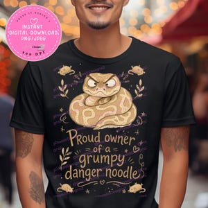 May include: A black t-shirt featuring a graphic illustration of a coiled, grumpy-looking snake in shades of tan and brown. The text reads "Proud owner of a grumpy danger noodle" in a stylized font, surrounded by small mouse and leaf illustrations.