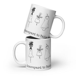 May include: Two white ceramic mugs stacked vertically, each featuring a black line drawing of three ghost-like vegetables including a carrot, a radish, and an eggplant. The text "Graveyard to Table" is printed in a typewriter-style font.