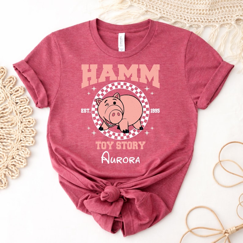 Może przedstawiać: Ciemnor&oacute;żowy melanżowy t-shirt z kr&oacute;tkim rękawem i grafiką przedstawiającą postać Hamma z Toy Story. Wz&oacute;r zawiera okrąg w szachownicę, napis HAMM, EST. 1995, TOY STORY oraz imię Aurora wypisane białą czcionką pisankową poniżej.