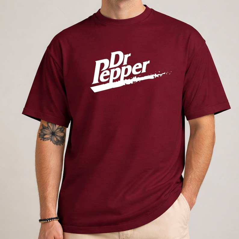 May include: A maroon t-shirt featuring the Dr Pepper logo in white. The logo includes the words "Dr Pepper" in a stylized font with a white streak underneath. The shirt has short sleeves and a crew neck.