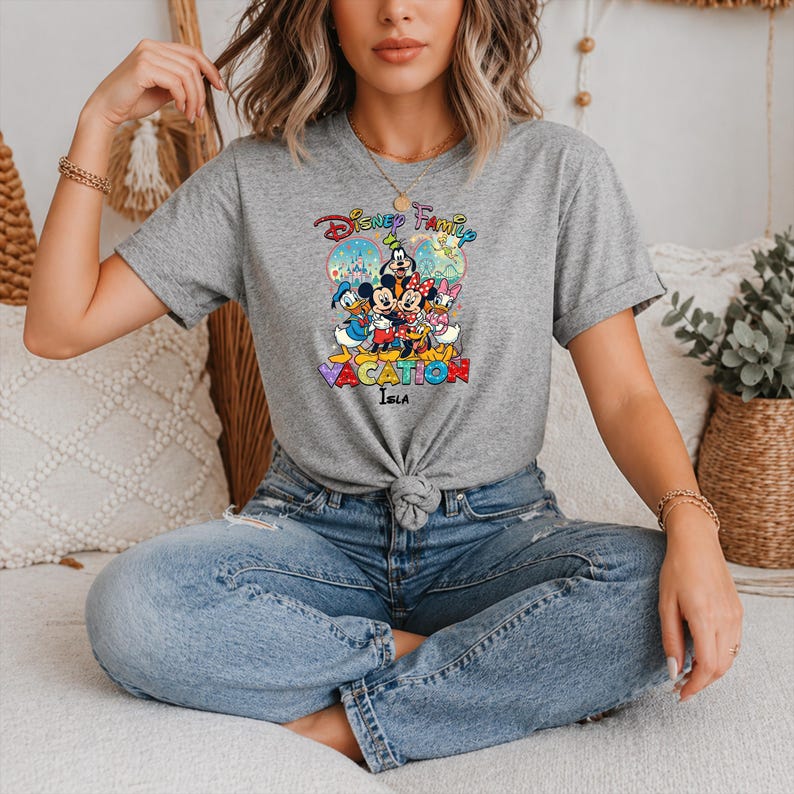 May include: A gray t-shirt featuring a colorful graphic of Disney characters, including Mickey Mouse, Minnie Mouse, Donald Duck, and Daisy Duck. The shirt has the words "Disney Family Vacation" printed on it. The shirt is knotted at the waist.