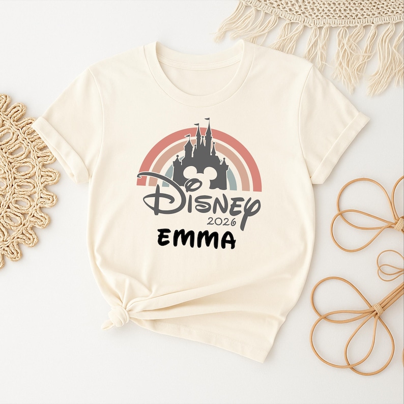 May include: A cream-coloured t-shirt with a Disney castle design set against a rainbow. The design includes the word "Disney", the year "2026", and the name "EMMA" in black. The shirt has short sleeves and a relaxed fit.