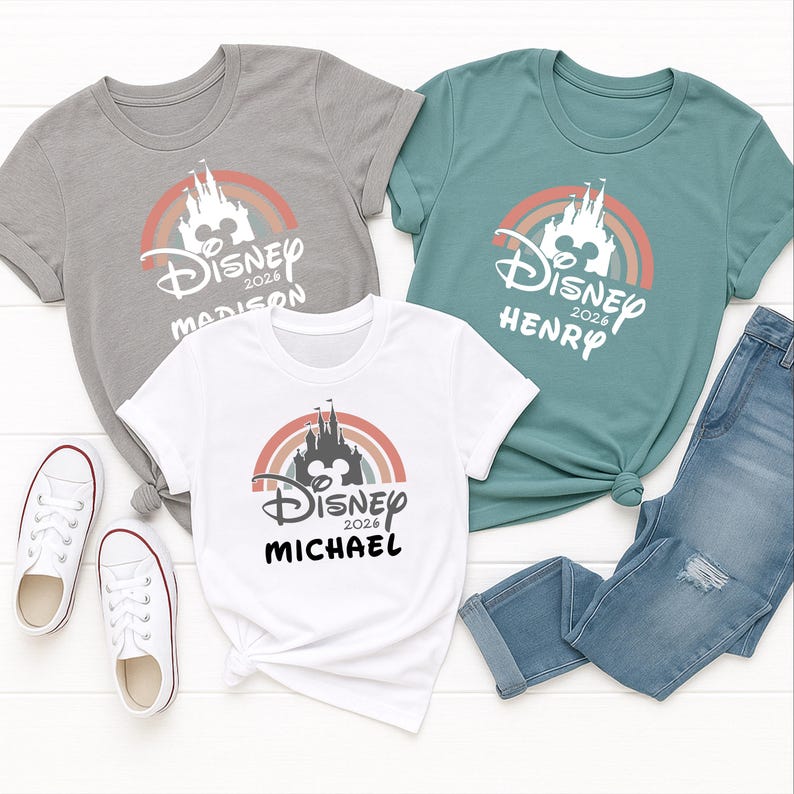 May include: Three t-shirts featuring a Disney castle and rainbow design. The shirts are in grey, teal, and white, with names and the year 2026 printed below the logo. A pair of blue jeans and white trainers are also in the image.