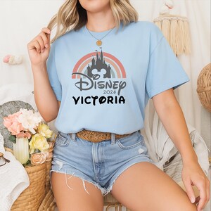 May include: Pale blue t-shirt with a rainbow design, a castle silhouette, and the text "Disney 2026 Victoria". The top has short sleeves and a round neckline.