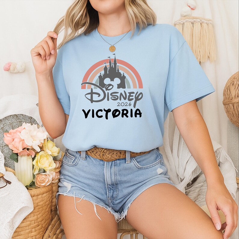 May include: Pale blue t-shirt with a rainbow design, a castle silhouette, and the text "Disney 2026 Victoria". The top has short sleeves and a round neckline.