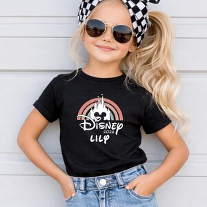 May include: Black t-shirt with a Disney castle design, rainbow, and the text "Disney 2026 Lily". A young person wears the shirt, along with sunglasses and a black and white checkered headband.
