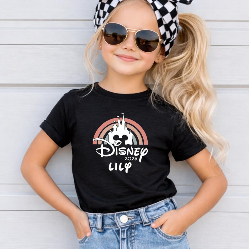 May include: Black t-shirt with a Disney castle design, rainbow, and the text "Disney 2026 Lily". A young person wears the shirt, along with sunglasses and a black and white checkered headband.
